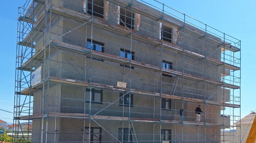 Építési fázisban lévő bérház állványzattal, látható héjjal, külső falazattal, kék ég a háttérben.