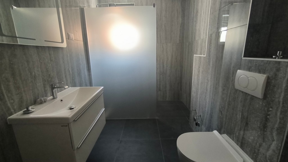 Modern kialakítású zuhanyzós fürdőszoba, horvátországi ingatlan Simuniban, szürke csempe és erős fény kiemelve.