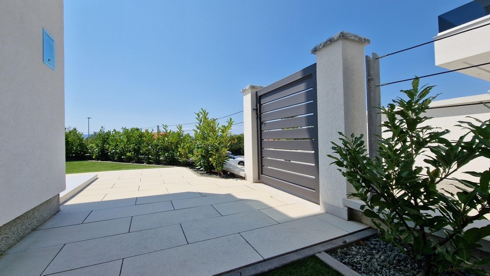 Modern épület bejárati kapuja burkolt előtérrel és zöld növényekkel.