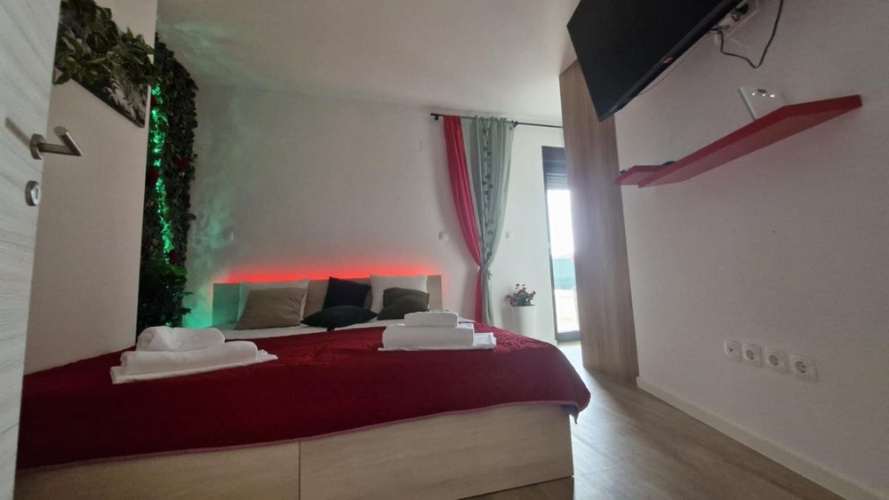 Hálószoba piros ágytakaróval, dekoratív háttérrel és TV-vel a falon