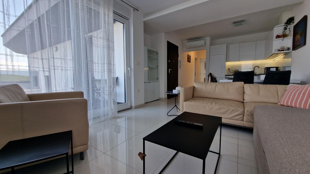 Nyitott nappali világos bútorokkal, üvegajtó a teraszra és beépített konyhasarok.