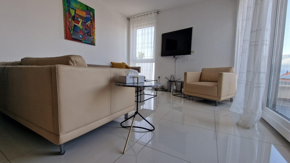 Stílusosan berendezett nappali kanapéval, televízióval és nagy ablakokkal, amelyek sok fényt engednek be.