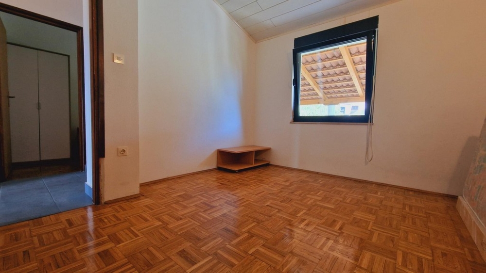 Üres hálószoba nagy ablakkal, egyszerű bútorokkal és fapadlóval.