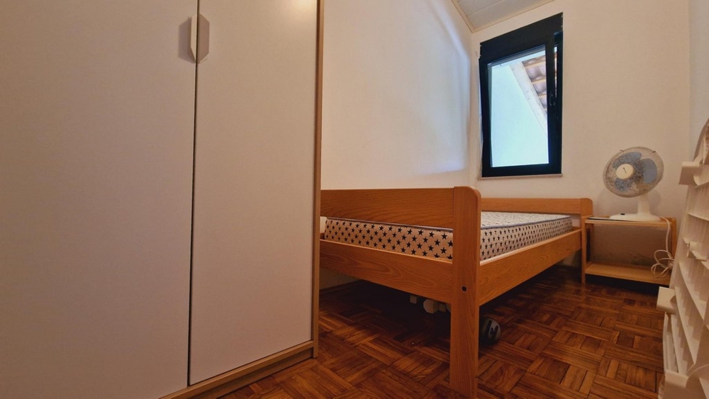 Kis hálószoba ággyal, szekrénnyel és ablakkal, egyszerű berendezéssel.