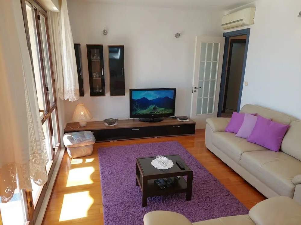 Nappali kanapéval, televízióval, lila párnákkal és szőnyeggel, nagy ablakokkal