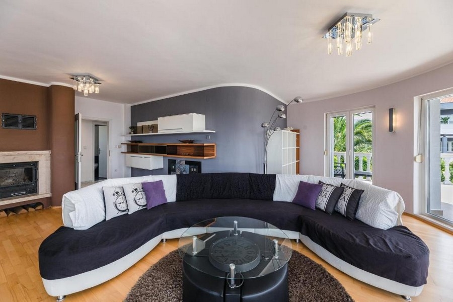Elegáns nappali egy horvátországi apartmanházban, tengerre néző kilátással, nagy íves kanapéval és kerek üvegasztallal barna szőnyegen, kandallóval és nyitott erkéllyel.