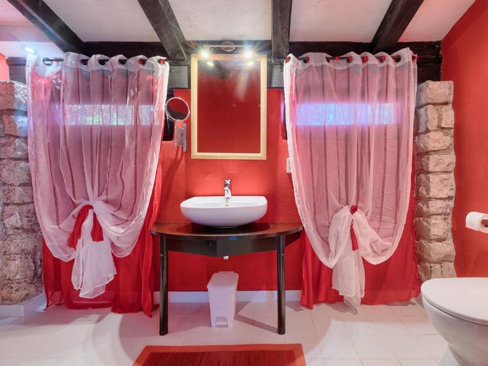Egy horvátországi ingatlan fürdőszobája piros falakkal, fehér vízvezetékkel, rusztikus asztalon mosdóval és átlátszó piros függönyökkel az ablakokon