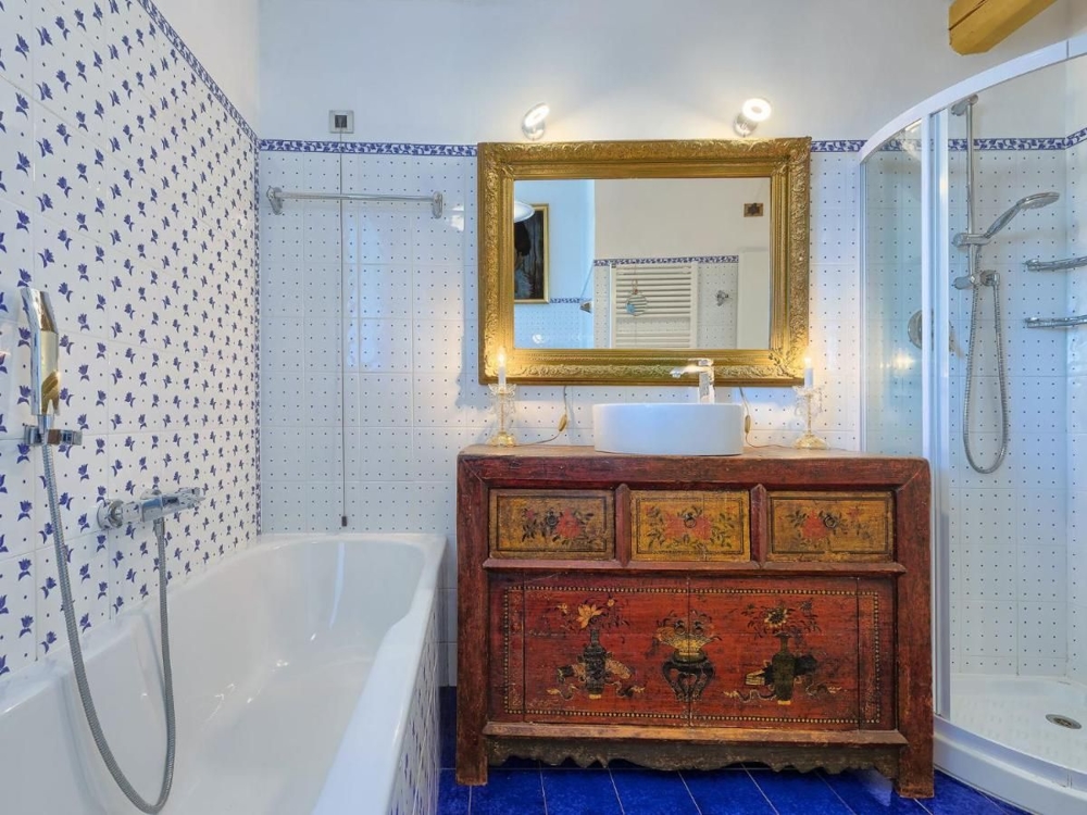 Kék-fehér csempével díszített fürdőszoba egy horvát ingatlanban, fürdőkáddal, zuhanyzóval és egy arany tükör alatti antik színes szekrénnyel