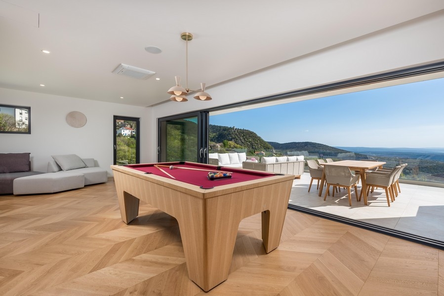 Világos és modern játékszoba, amelynek közepén egy piros fedett biliárdasztal, elegáns fapadló és a padlótól a mennyezetig érő ablakokon keresztül lélegzetelállító kilátás nyílik a dombokra.