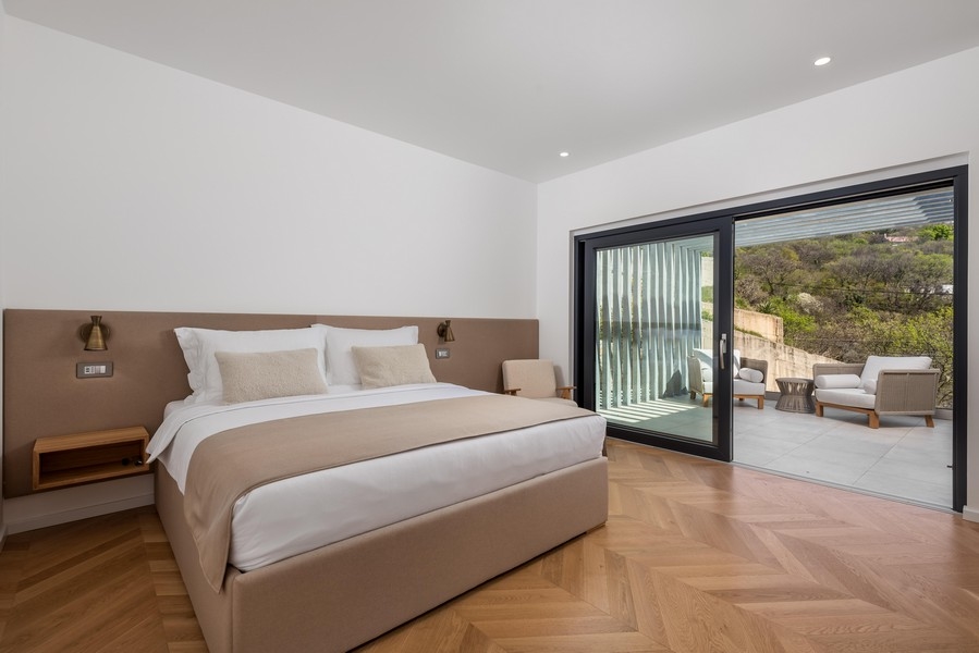 Minimalista hálószoba nagy franciaággyal, bézs színű ágyneművel és fejtámlával, fapadlóval és tolóajtóval, amely egy ülősarokkal és gyönyörű kilátással rendelkező teraszra vezet.