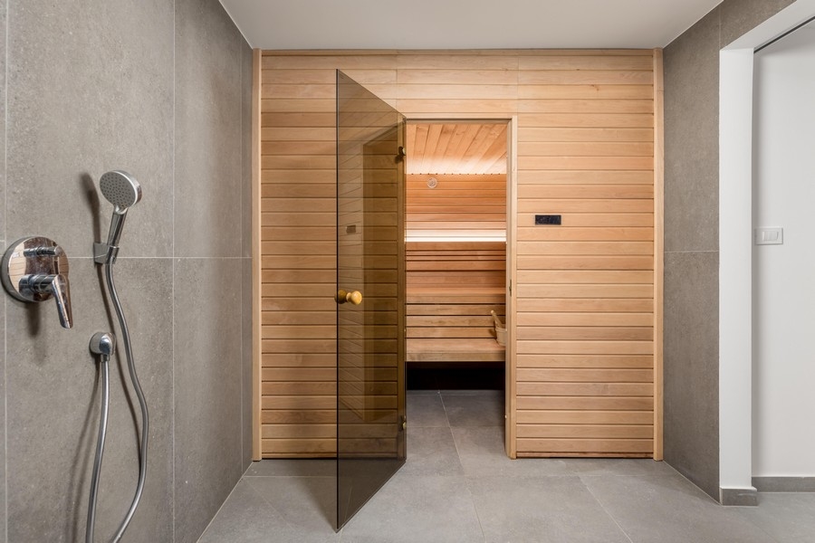A bal oldalon egy modern zuhanyzós fürdőszoba, a jobb oldalon egy fa szauna. A falakat szürke csempék borítják, és üvegajtó vezet a szaunába.