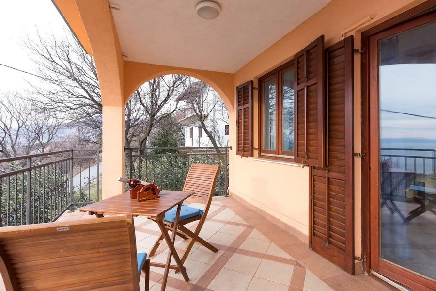 Fedett erkély fa székekkel és asztallal, kilátással a lombos környékre és a távolban a tengerre.