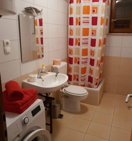 Funkcionális fürdőszoba zuhanyfüggönnyel, mosógéppel és piros és narancssárga kiegészítőkkel, mosdóval, WC-vel és kis ablakkal.