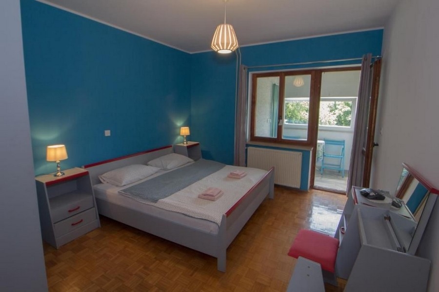 Egy hálószoba kékre festett falakkal, egy nagy franciaággyal, két éjjeliszekrény lámpákkal, egy ablak és egy ajtó, amely az erkélyre vezet.