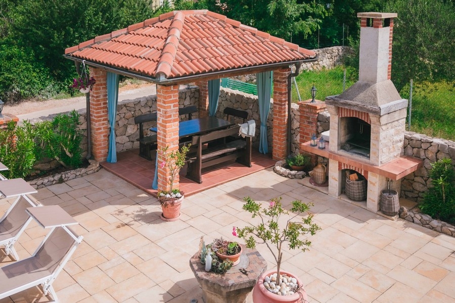 Kültéri grillező tégla pavilonnal, fa étkezőasztallal és kőkemencével a csempézett teraszon