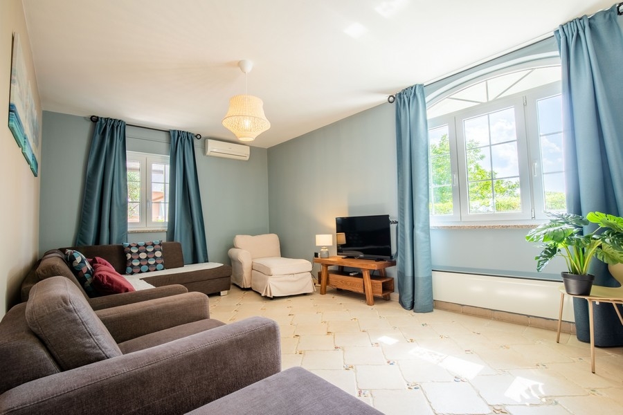 Hangulatos nappali több kanapéval, TV-vel és nagy ablakokkal, kék függönyökkel, amelyek beengedik a természetes fényt