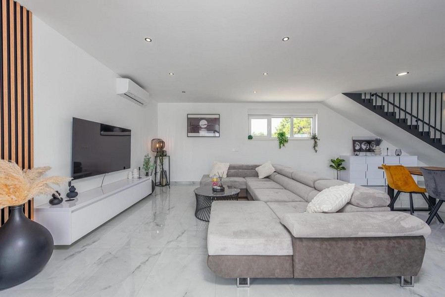 Nyitott nappali nagy szürke kanapéval, fehér falakkal, kilátással az étkezőre és a lépcsőkre.
